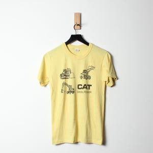 Puede incluir: Camiseta amarilla clara con un gr&aacute;fico negro de veh&iacute;culos de construcci&oacute;n y el texto "CAT DIESEL POWER". La camiseta est&aacute; colgada en una percha negra sobre un fondo blanco.
