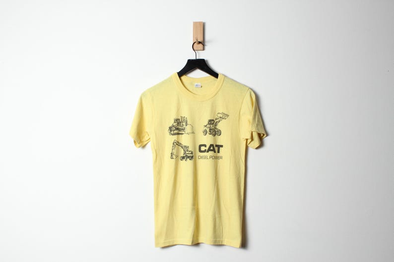 Puede incluir: Camiseta amarilla clara con un gr&aacute;fico negro de veh&iacute;culos de construcci&oacute;n y el texto "CAT DIESEL POWER". La camiseta est&aacute; colgada en una percha negra sobre un fondo blanco.