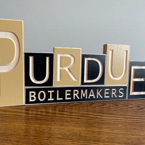 May include: Wooden block letters spelling "Purdue Boilermakers" in gold and black paint.