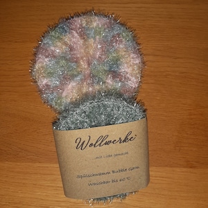 Könnte beinhalten: Ein grüner und rosa gestrickter Spülschwamm aus Bubblegarn. Der Schwamm ist mit "Wollwerke... mit Liebe gemacht" und "Spülschwamm Bubble Garn Waschbar bis 60°C" beschriftet.