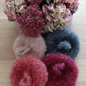 Könnte beinhalten: Vier gehäkelte Schwämme in verschiedenen Farben: rosa, rot, burgunderrot und grau. Die Schwämme sind aus einem flauschigen, glitzernden Garn gefertigt.