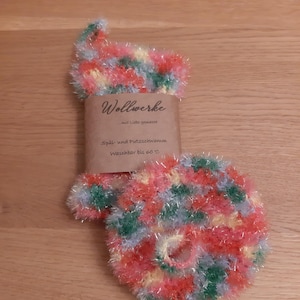 Könnte beinhalten: Ein farbenfroher, handgestrickter Spülschwamm mit einer Schlaufe. Der Schwamm besteht aus Garn in den Farben Rot, Gelb, Grün und Blau. Ein braunes Papieretikett ist mit dem Text "Wollwerke" befestigt.