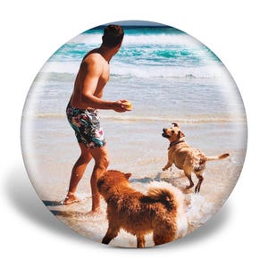 Op de afbeelding: Een persoon in een zwembroek met bloemenprint op een strand, op het punt om een bal te gooien voor twee honden. E&eacute;n hond rent naar de bal, de andere is in het water. De oceaan is op de achtergrond.