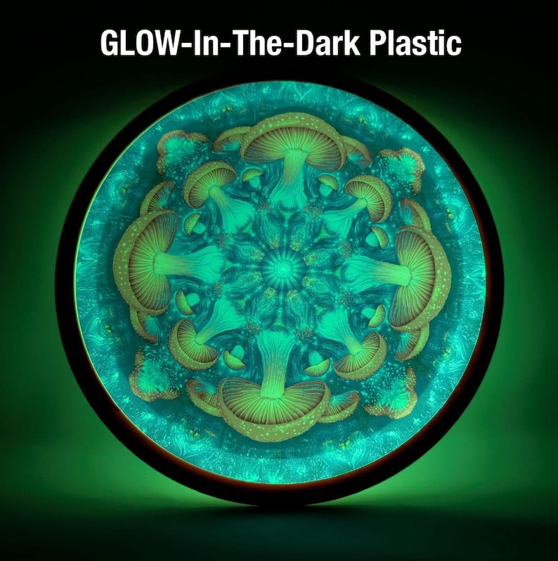 Op de afbeelding: Een ronde, oplichtende plastic schijf met een symmetrisch ontwerp van groene paddenstoelen. De paddenstoelen zijn gerangschikt in een radiaal patroon tegen een turquoise achtergrond. De tekst "GLOW-In-The-Dark Plastic" staat bovenaan.