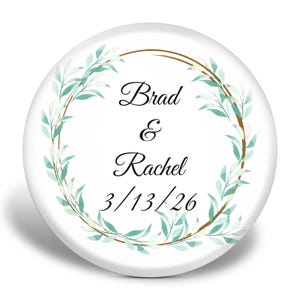 May include: A white, circular button featuring the names "Brad & Rachel" and the date "3/13/26" in a stylish script. The text is encircled by a gold ring and a wreath of green leaves.
