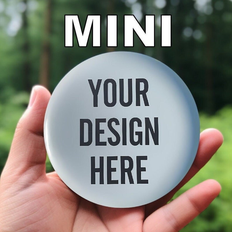 以下が含まれることがあります： 手で持たれた、黒い文字で「YOUR DESIGN HERE」と書かれた丸い水色のボタン。ボタンの上には白い文字で「MINI」と書かれています。背景はぼやけた緑色です。