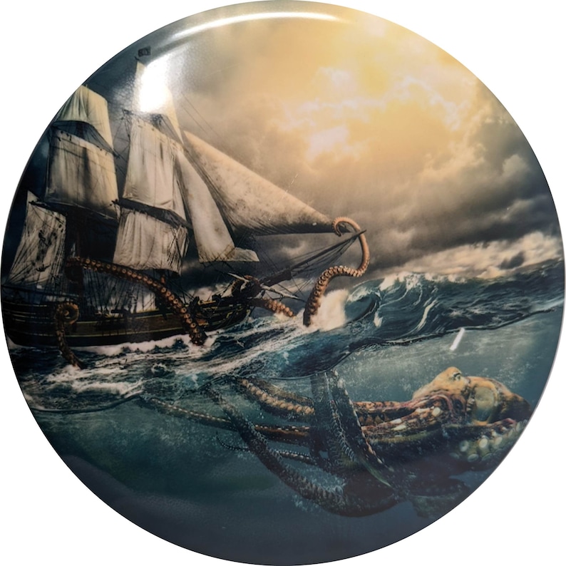 Op de afbeelding: Ronde decoratieve plaat met een dramatische nautische sc&egrave;ne. Een groot schip wordt aangevallen door een gigantische octopus in woelige wateren onder een stormachtige hemel. Het kunstwerk is gedetailleerd met tinten blauw, bruin en wit.