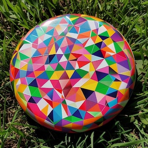 Op de afbeelding: Een rond, kleurrijk object met een geometrisch patroon van driehoeken in verschillende kleuren, waaronder roze, geel, groen, blauw en oranje. Het object ligt op groen gras.