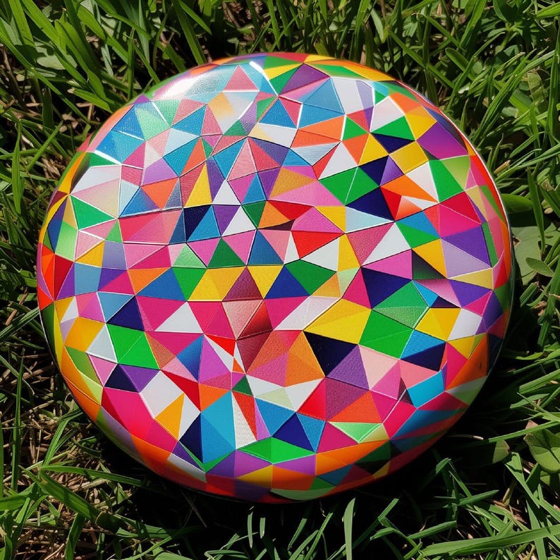 Op de afbeelding: Een rond, kleurrijk object met een geometrisch patroon van driehoeken in verschillende kleuren, waaronder roze, geel, groen, blauw en oranje. Het object ligt op groen gras.