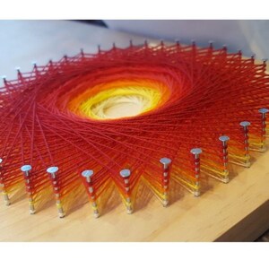 Pode incluir: Uma pe&ccedil;a circular de arte de corda com um degradado de fio vermelho, laranja e amarelo enrolado em torno de pregos martelado em uma t&aacute;bua de madeira.