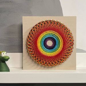Pode incluir: Uma pe&ccedil;a de arte de cordas com cores do arco-&iacute;ris sobre um fundo de madeira. A arte de cordas tem a forma de um c&iacute;rculo com um centro de cor roxo escuro. A arte de cordas &eacute; cercada por uma moldura de madeira clara.
