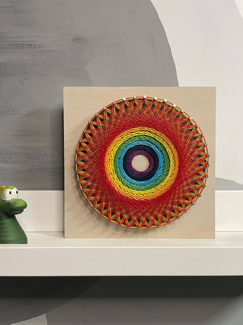 Pode incluir: Uma pe&ccedil;a de arte de cordas com cores do arco-&iacute;ris sobre um fundo de madeira. A arte de cordas tem a forma de um c&iacute;rculo com um centro de cor roxo escuro. A arte de cordas &eacute; cercada por uma moldura de madeira clara.