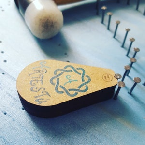Puede incluir: Un tablero de madera para manualidades con un diseño azul y blanco. El tablero tiene forma de lágrima y tiene las palabras "Craft Stuff" escritas en él. Hay varios clavos metálicos que sobresalen del tablero.