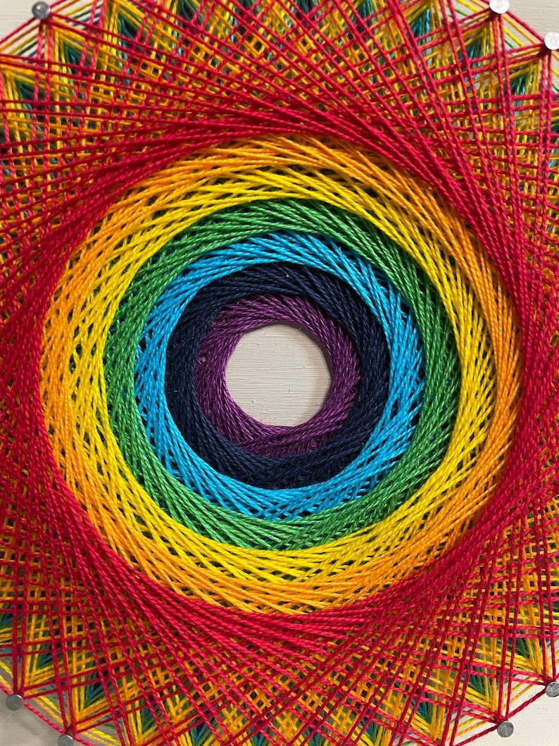 Pode incluir: Um design de arte de barbante arco-&iacute;ris com um padr&atilde;o circular. A arte de barbante &eacute; feita com barbante colorido e pregos martelado em uma t&aacute;bua de madeira.