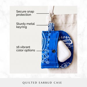 May include: A blue and white paisley quilted earbud case featuring a secure snap closure, a sturdy metal keyring, and text labels indicating secure snap protection, sturdy metal keyring, and 16 vibrant color options. The case holds white earbuds.