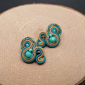 Może przedstawiać: Para ręcznie robionych kolczyków z wzorem soutache. Kolczyki mają turkusowy i beżowy wzór przypominający linę z turkusowymi koralikami. Kolczyki są umieszczone na drewnianym tle.