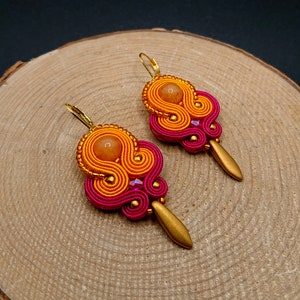 Pode incluir: Um par de brincos pendentes com um design rosa e laranja. Os brincos têm um gancho de metal dourado e uma pequena pedra redonda laranja no centro.