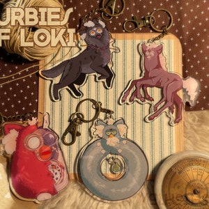 May include: Keychains featuring cartoon-style illustrations of Furby-like creatures in various forms. The keychains include a red Furby, a black dog, a purple horse, and a blue Furby in a life preserver. The text "FURBIES OF LOKI" is visible.