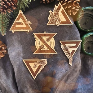 May include: A collection of six wooden laser-cut geometric shapes, including triangles and a stylized hourglass. The shapes feature intricate designs and patterns, with a natural wood grain finish. The items are arranged on a gray fabric surface.