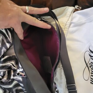 May include: A black and white zebra print tote bag with a purple lining and a silver zipper. The bag has a black adjustable strap and a silver heart-shaped logo. The bag is shown with a white dust bag with the word "Brighton" printed on it.