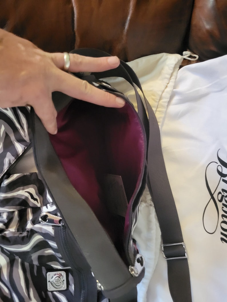 May include: A black and white zebra print tote bag with a purple lining and a silver zipper. The bag has a black adjustable strap and a silver heart-shaped logo. The bag is shown with a white dust bag with the word "Brighton" printed on it.
