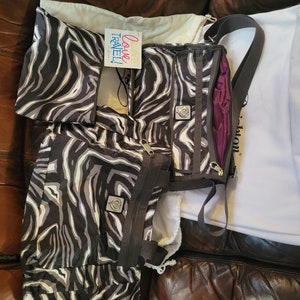 May include: A black and white zebra print tote bag with a zipper closure and a small silver logo. The bag has a long strap and is sitting on a brown leather couch. The text "Live Love Travel!" is visible on a small tag attached to the bag.