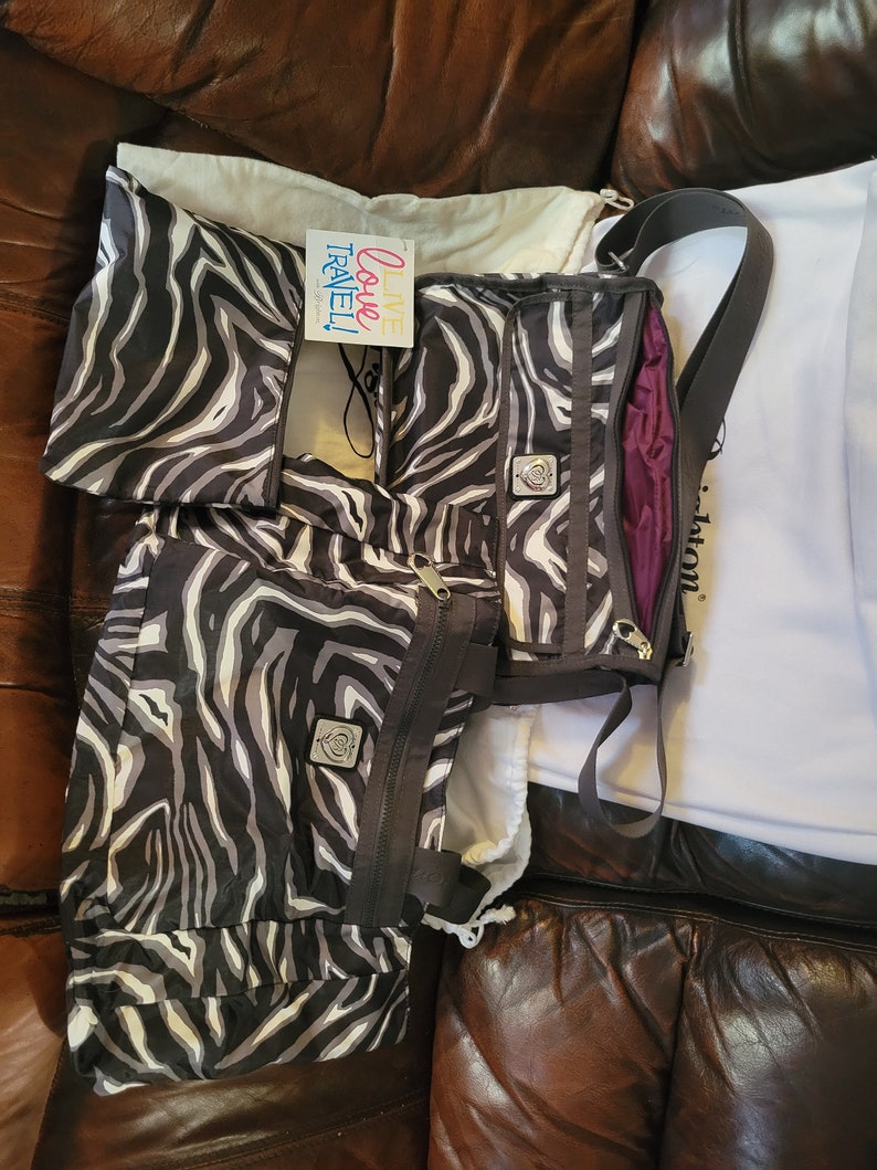 May include: A black and white zebra print tote bag with a zipper closure and a small silver logo. The bag has a long strap and is sitting on a brown leather couch. The text "Live Love Travel!" is visible on a small tag attached to the bag.