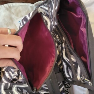 May include: A black and white zebra print tote bag with a pink interior. The bag has a zipper closure and a small pocket on the front. The bag has a black strap and the word "Brighton" is visible on the side.