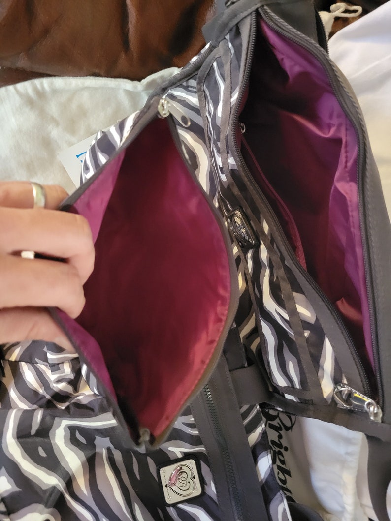 May include: A black and white zebra print tote bag with a pink interior. The bag has a zipper closure and a small pocket on the front. The bag has a black strap and the word "Brighton" is visible on the side.