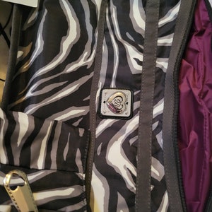 May include: A black and white zebra print crossbody bag with a silver heart-shaped logo and a purple lining. The bag has a zipper closure and a strap.