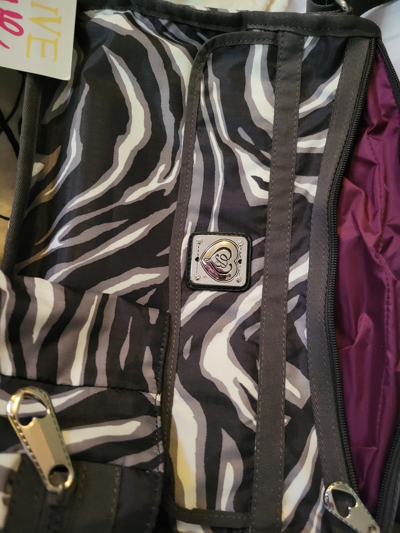 May include: A black and white zebra print crossbody bag with a silver heart-shaped logo and a purple lining. The bag has a zipper closure and a strap.
