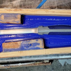Op de afbeelding: Een zilveren klauwhamer met een grijze en zwarte handgreep rust in een blauwe mal in een houten frame. Twee kleine houten blokken bevinden zich ook in de mal. De hamer is een gereedschap voor constructie en reparatie.