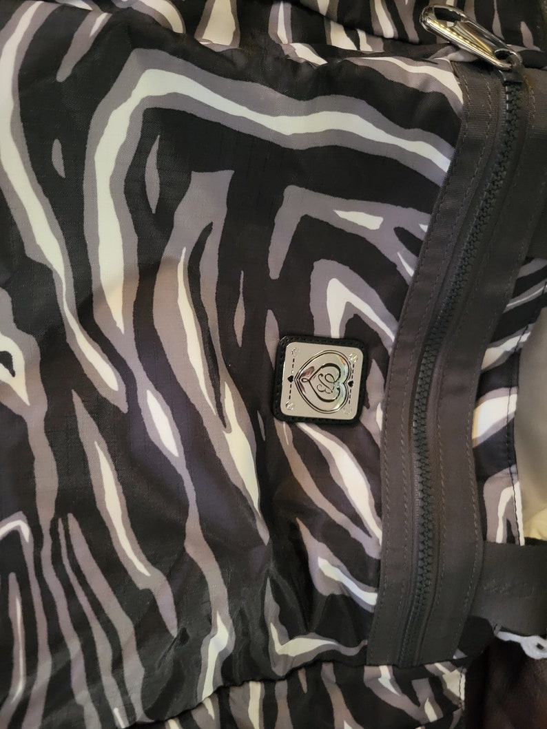 May include: A black and white zebra print tote bag with a silver heart logo. The bag has a zipper closure and a top handle.