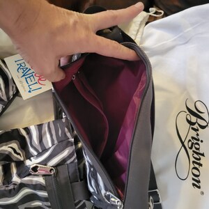 May include: A black and white zebra print crossbody bag with a zipper closure and a silver zipper pull. The bag has a burgundy lining and a black strap. The bag is made by Brighton.