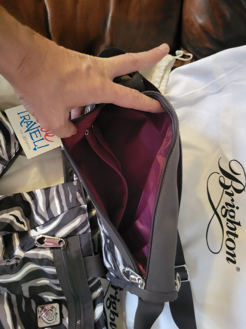 May include: A black and white zebra print crossbody bag with a zipper closure and a silver zipper pull. The bag has a burgundy lining and a black strap. The bag is made by Brighton.