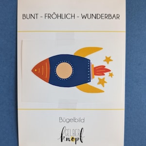 Könnte beinhalten: Ein farbenfroher Bügelbild mit einer Rakete mit gelben Flammen und Sternen. Der Text "BUNT - FRÖHLICH - WUNDERBAR" befindet sich oben auf dem Patch. Der Text "Bügelbild GELBER Knopf" befindet sich unten auf dem Patch.