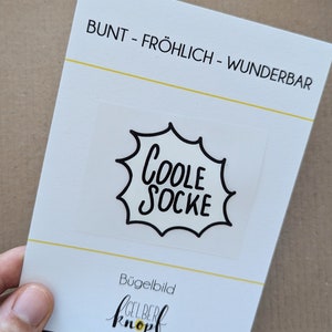 Könnte beinhalten: Ein weißer Bügelbild-Transfer mit schwarzem Text, der "Coole Socke" in einer Sprechblase zeigt. Der Transfer ist mit "Bügelbild" und "gelber Knopf" beschriftet.