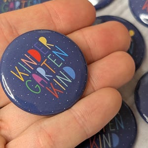 Könnte beinhalten: Eine Sammlung von marineblauen runden Knöpfen mit dem Schriftzug "KINDERGARTEN KIND" in bunten Buchstaben. Die Knöpfe haben eine glänzende Oberfläche und kleine weiße Punkte. Ein lustiges Accessoire für Schüler.