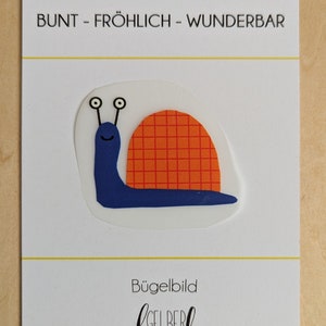 Könnte beinhalten: Eine blaue Schnecke mit einem rot-orangefarbenen karierten Haus. Die Schnecke hat zwei weiße Augen und einen lächelnden Mund. Das Bild ist auf einer weißen Karte mit dem Text "BUNT - FRÖHLICH - WUNDERBAR" und "Bügelbild GELBER Knopf".