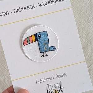 Könnte beinhalten: Ein weißer runder Bügelaufkleber mit einem blauen Tukan mit einem gelben, rosa und grünen Schnabel. Der Aufnäher trägt den Text "bunt - fröhlich - wunderbar" und "Aufnäher / Patch" und "gelber knopl" in schwarzer Schrift.
