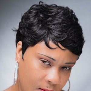 Peut inclure: Portrait rapproché d'une personne aux cheveux courts, noirs et bouclés. Elle porte des boucles d'oreilles en argent et a un piercing au nez. La personne regarde vers le bas et a une expression sérieuse.