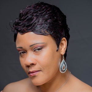 May include: A woman with short, dark hair wearing a light pink off-the-shoulder top. She is wearing a silver necklace with a heart pendant and silver teardrop earrings.