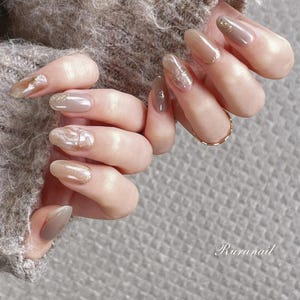 May include: Close-up of a hand with long, oval nails. The nails are painted in neutral shades of beige and taupe, with gold glitter accents. A gold ring adorns one finger. The image is set against a soft, neutral background. The word "Rurunail" is visible.