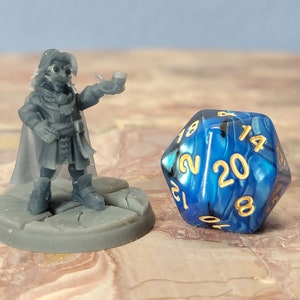 May include: A blue and black 20-sided die with gold numbers, next to a gray miniature figurine of a person holding a pipe.