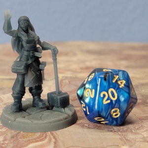 May include: A gray 3D printed miniature of a female figure holding a hammer, standing on a gray base. The figure is wearing a long tunic and has long hair. A blue and black 20-sided die with gold numbers is in the background.