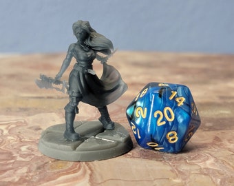 May include: A gray plastic miniature figurine of a female warrior holding a sword, standing on a gray base. The figurine is next to a blue and black 20-sided die with gold numbers.