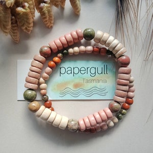 May include: A double-strand bracelet with a mix of wooden and stone beads in pink, beige, orange, and green. The bracelet features round and cylindrical beads. A card with the text "papergull Tasmania" is visible. The bracelet is a unique accessory.