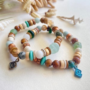 May include: Two beaded bracelets featuring a mix of natural wood, white, turquoise, and green beads. The bracelets include smooth round beads, flat disc-shaped spacers, and a small metal charm on each, one bronze and one blue, shaped like a spiral.