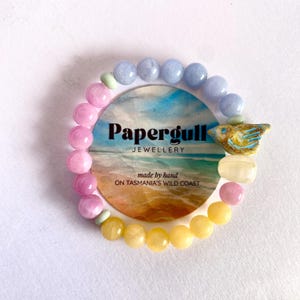 May include: A beaded bracelet featuring a circular display card with the text Papergull JEWELLERY made by hand ON TASMANIA'S WILD COAST. The bracelet consists of light blue, pink, yellow, and translucent beads, with a ceramic bird charm.