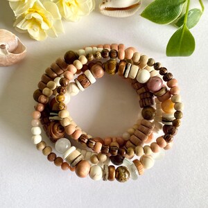 May include: A stack of multiple beaded bracelets featuring a variety of round and cylindrical beads in shades of tan, brown, cream, and soft pink. The beads are crafted from wood, stone, and smooth polished materials, arranged in a circular pattern.